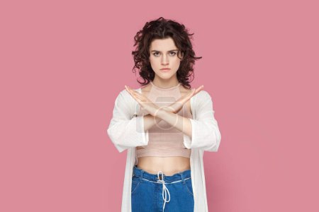 Photo for Portrait of serious woman with curly hair wearing casual style outfit crossed her arms, showing no way gesture, looking at camera. Indoor studio shot isolated on pink background. - Royalty Free Image