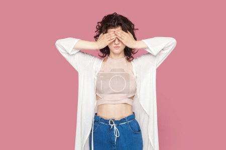 Photo for Portrait of scared young adult woman with curly hair wearing casual style outfit covering her eyes with palms, doesn't want to see something shameful. Indoor studio shot isolated on pink background. - Royalty Free Image