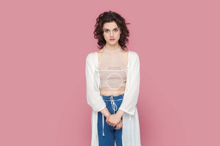 Photo for Portrait of serious concentrated woman with curly hair wearing casual style outfit standing looking at camera with attentive expression. Indoor studio shot isolated on pink background. - Royalty Free Image