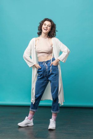 Photo for Full length of beautiful brunette woman in casual style attire standing and looking at camera with happy face, laughing, keeps hands on hips. Indoor studio shot isolated on blue background. - Royalty Free Image