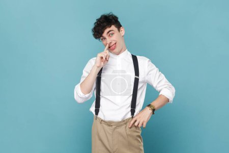 Photo for Portrait of mischievous man in shirt and suspender putting finger into his nose and showing tongue, fooling around, bad habits, disrespectful behavior. Indoor studio shot isolated on blue background. - Royalty Free Image
