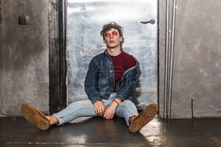 Photo for Portrait of young self-confident fashion model man in bright red sunglasses and denim casual style, sitting on floor near metallic door, looking at camera with relaxed look. Indoor studio shot. - Royalty Free Image