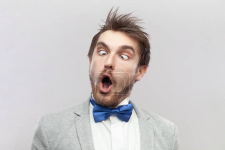 Photo for Portrait of funny crazy childish bearded man keeps mouth open and eyes crossed, making foolish face, wearing grey suit and blue bow tie. Indoor studio shot isolated on gray background. - Royalty Free Image
