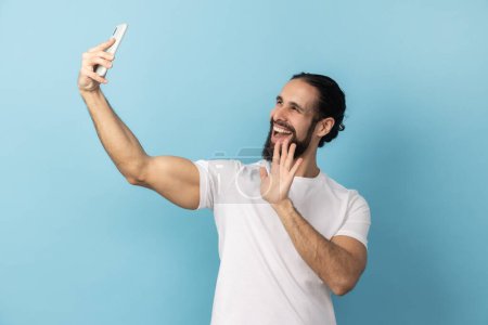 Téléchargez les photos : Portrait d'homme barbu portant un T-shirt blanc prenant un selfie et agitant la main, faisant signe de bonjour aux disciples, communiquant sur appel vidéo. Studio intérieur tourné isolé sur fond bleu. - en image libre de droit