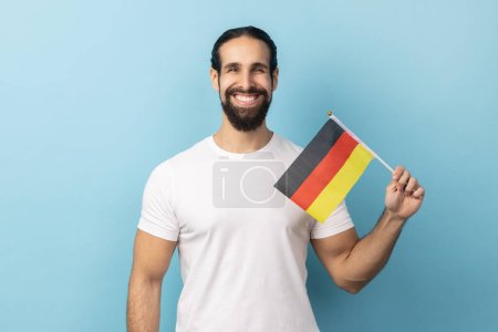 Téléchargez les photos : Portrait d'un homme souriant avec une barbe portant un T-shirt blanc tenant le drapeau de l'Allemagne, célébrant le Jour de l'Allemagne - 3 octobre, regardant la caméra. Studio intérieur tourné isolé sur fond bleu. - en image libre de droit