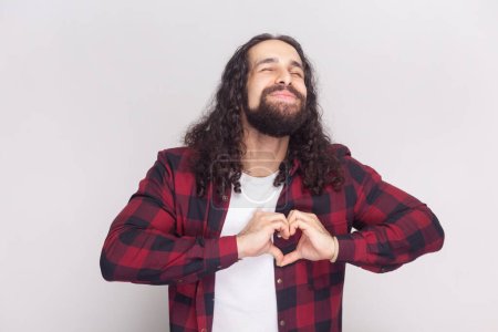Photo for Pleased good looking man in checkered red shirt shapes heart over chest, confesses in love, has romantic mood, expresses truthful feelings. Indoor studio shot isolated on gray background. - Royalty Free Image
