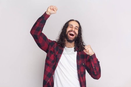 Photo for Cheerful triumphing man with long curly hair in checkered red shirt achieves victory, raises clenched fists with triumph, rejoices winning prize. Indoor studio shot isolated on gray background. - Royalty Free Image