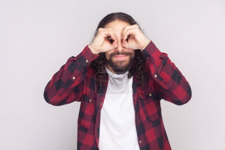 Photo for Portrait of bearded man with long curly hair in checkered red shirt making glasses shape with fingers, looking through binoculars hand gesture. Indoor studio shot isolated on gray background. - Royalty Free Image