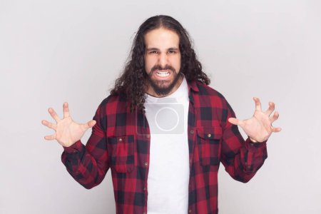 Photo for Portrait of annoyed bearded man with long curly hair in checkered red shirt gestures with anger, screams at someone, feels outraged frowns face. Indoor studio shot isolated on gray background. - Royalty Free Image
