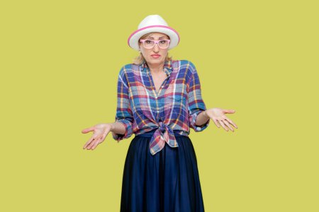 Photo for Puzzled clueless senior woman wearing checkered shirt, hat and eyeglasses receives strange offer, shrugs shoulders with hesitation, feels doubt. Indoor studio shot isolated on yellow background. - Royalty Free Image