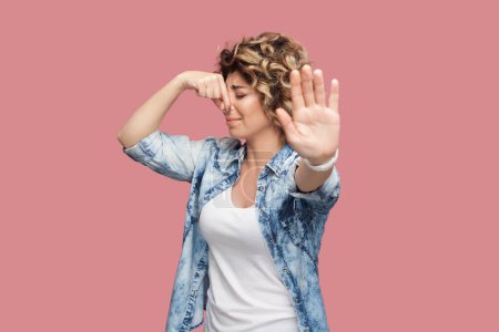 Photo for Portrait of attractive woman with curly hairstyle wearing blue shirt standing pinching nose, bad smell, turning away, showing stop gesture. Indoor studio shot isolated on pink background. - Royalty Free Image