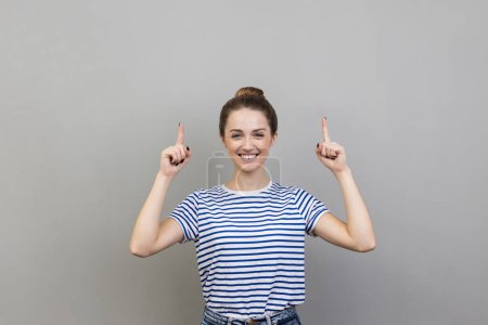 Téléchargez les photos : Portrait d'une femme séduisante extrêmement heureuse portant un T-shirt rayé pointant les doigts vers le haut sur un espace vide, satisfaite de la zone publicitaire. Studio intérieur tourné isolé sur fond gris. - en image libre de droit