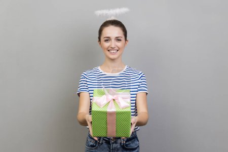 Porträt einer lächelnden zufriedenen Frau mit gestreiftem T-Shirt und Nimbus über dem Kopf, die grüne Geschenkschachtel überreicht und mit Urlaub gratuliert. Indoor Studio isoliert auf grauem Hintergrund gedreht.