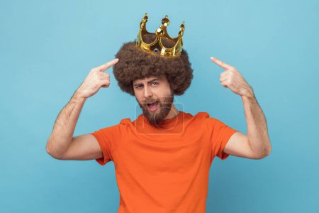 Portrait of self confident man with Afro hairstyle wearing orange T-shirt standing and pointing at golden crown, looking at camera with proud expression. Indoor studio shot isolated on blue background