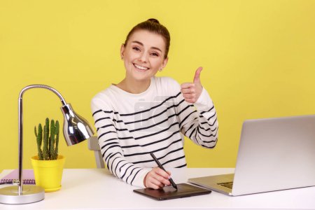 Photo for Satisfied smiling woman creative designer sitting at workplace with graphic tablet and laptop, looking at camera and showing thumb up. Indoor studio studio shot isolated on yellow background. - Royalty Free Image