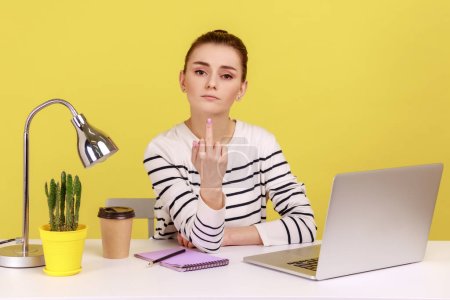 Uncultured aggressive woman office manager sitting at workplace and showing middle finger fuck sign and looking at camera with serious face. Indoor studio studio shot isolated on yellow background.