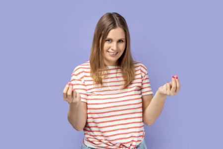 Photo for Portrait of smiling satisfied beautiful blond woman wearing striped T-shirt standing and showing money gesture with hands, looking at camera. Indoor studio shot isolated on purple background. - Royalty Free Image