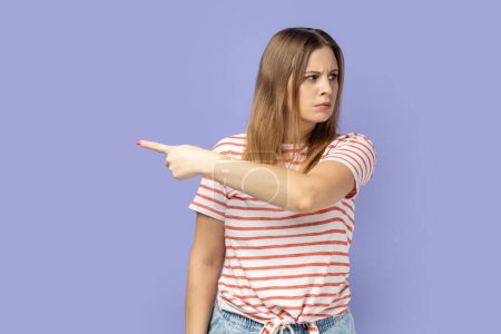Photo for Upset angry blond woman wearing striped T-shirt pointing finger away looking with resentful expression, confused with conflict, kicking out. Indoor studio shot isolated on purple background. - Royalty Free Image