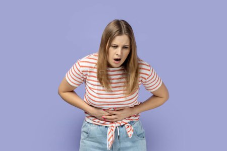 Photo for Period pain. Portrait of blond woman wearing striped T-shirt standing and feeling pain on her stomach, holding hands on belly, frowning face. Indoor studio shot isolated on purple background. - Royalty Free Image