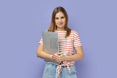 Photo for Portrait of cheerful happy optimistic blond woman wearing striped T-shirt standing with closed laptop, finishing her work, being satisfied. Indoor studio shot isolated on purple background. - Royalty Free Image