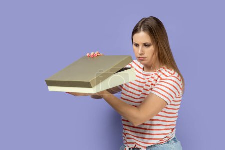 Photo for Portrait of sad blond woman wearing striped T-shirt standing holding present box and looking inside and expressing disappointment of gift. Indoor studio shot isolated on purple background. - Royalty Free Image