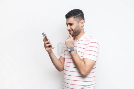 Téléchargez les photos : Portrait d'un homme barbu heureux et souriant portant un t-shirt rayé debout à l'aide d'un téléphone portable, regardant une vidéo ou vérifiant les réseaux sociaux. Studio intérieur tourné isolé sur fond gris. - en image libre de droit