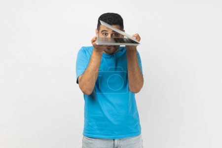 Téléchargez les photos : Portrait d'un homme drôle positif non rasé portant un T-shirt bleu debout regardant la caméra à travers un ordinateur portable à moitié ouvert, regardant. Studio intérieur tourné isolé sur fond gris. - en image libre de droit