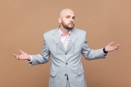 Photo for Portrait of hesitant puzzled bald bearded man shrugs shoulders in bewilderment, looks confusingly at camera, wearing gray jacket. Indoor studio shot isolated on brown background. - Royalty Free Image