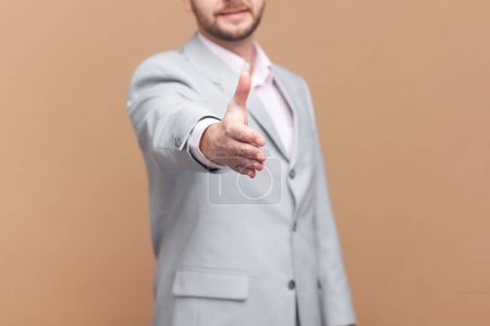 Photo for Portrait of unknown man stretching hand for handshaking, accepting job offer, partnership, beginning or end of meeting, wearing gray jacket. Indoor studio shot isolated on brown background. - Royalty Free Image