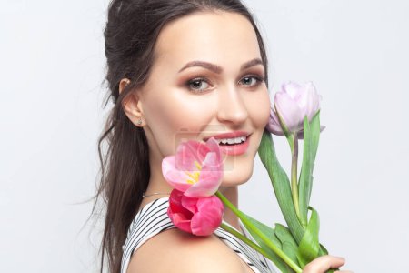 Photo for Side view portrait of smiling beautiful brunette woman holding bouquet of tulips, looking at camera, smiling with happiness, wearing striped dress. Indoor studio shot isolated on gray background. - Royalty Free Image