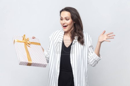 Photo for Portrait of amazed happy positive brunette woman gets present on her birthday, rejoicing, screaming with happiness, wearing striped jacket. Indoor studio shot isolated on gray background. - Royalty Free Image