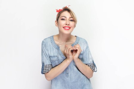Téléchargez les photos : Portrait d'une femme blonde romantique positive et joyeuse portant une chemise en denim bleu et un bandeau rouge montrant un geste de cœur, l'amour de l'expression. Studio intérieur tourné isolé sur fond gris. - en image libre de droit