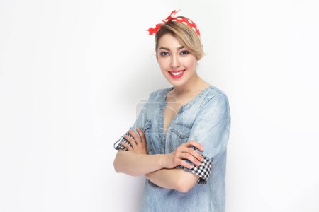 Photo for Portrait of happy joyful beautiful blonde woman wearing blue denim shirt and red headband standing with crossed arms, looking smiling at camera. Indoor studio shot isolated on gray background. - Royalty Free Image