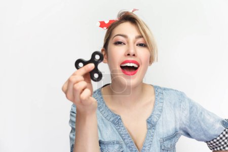 Photo for Portrait of excited amazed laughing blonde woman wearing blue denim shirt and red headband standing playing with spinner fidget. Indoor studio shot isolated on gray background. - Royalty Free Image