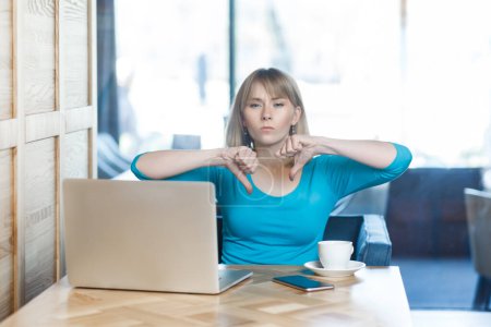 Photo for Portrait of serious attractive young woman with blonde hair in blue shirt working on laptop, showing thumbs down, negative feedback. Indoor shot in cafe with big window on background. - Royalty Free Image