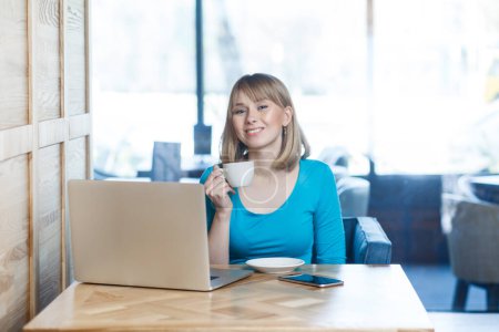 Téléchargez les photos : Portrait d'une jeune femme souriante positive aux cheveux blonds en chemise bleue travaillant sur ordinateur portable, faisant une pause, buvant du café chaud, appréciant son travail. Intérieur tourné dans un café avec une grande fenêtre sur fond. - en image libre de droit