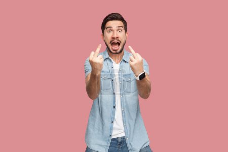 Portrait of bearded man in blue casual style shirt standing shows fuck you sign, being vulgar and has quarrel with someone, demonstrates middle finger. Indoor studio shot isolated on pink background.