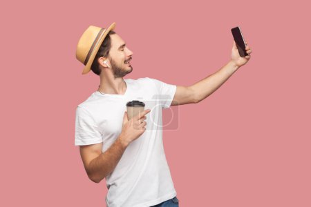 Foto de Retrato de vista lateral de hombre barbudo atractivo y sonriente en camiseta blanca y sombrero de pie sosteniendo café para llevar y haciendo selfie en el teléfono inteligente. Estudio interior plano aislado sobre fondo rosa. - Imagen libre de derechos