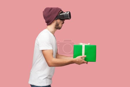 Photo for Surprised handsome man in white T-shirt and beany hat standing, holding green wrapped present box, giving gift, playing video game, posing in vr. Indoor studio shot isolated on pink background. - Royalty Free Image
