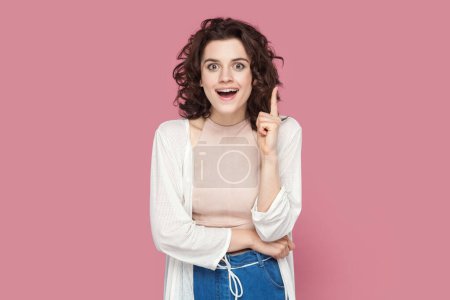Photo for Portrait of clever smart happy woman with curly hair wearing casual style outfit raises her index finger up, having good idea. Indoor studio shot isolated on pink background. - Royalty Free Image