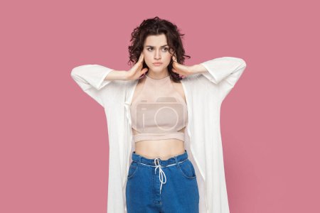Photo for Portrait of annoyed irritated woman with curly hair wearing casual style outfit covering ears with hands, looking away with frustrated facial expression. Indoor studio shot isolated on pink background. - Royalty Free Image