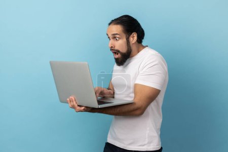Photo for Portrait of surprised man with beard entrepreneur, looking with open mouth at camera, achieving business goals, while working on computer laptop. Indoor studio shot isolated on blue background. - Royalty Free Image