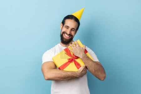 Photo for Portrait of joyful man with beard wearing white T-shirt and party cone holding embracing yellow wrapped present box, being satisfied to get gift. Indoor studio shot isolated on blue background. - Royalty Free Image