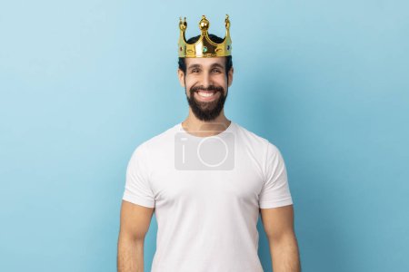 Photo for Portrait of man wearing white T-shirt and crown on head, smiling, concept of self confidence in success, self-motivation and dreams to be best. Indoor studio shot isolated on blue background. - Royalty Free Image