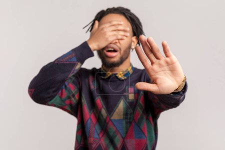 Photo for Portrait of scared shocked african-american man with dreadlocks and beard covering eyes with palm, showing stop gesture, sees something shameful. Indoor studio shot isolated on gray background. - Royalty Free Image