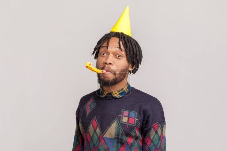 Photo for Portrait of positive funny african-american man in yellow festive cone with dreadlocks and beard blowing party horn, looking at camera. Indoor studio shot isolated on gray background. - Royalty Free Image