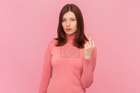 Unhöfliche Frau mit braunen Haaren, die den Mittelfinger zeigt und bittet, auszusteigen, Blick in die Kamera, respektloses Verhalten, trägt rosa Rollkragen. Indoor-Studio isoliert auf rosa Hintergrund aufgenommen