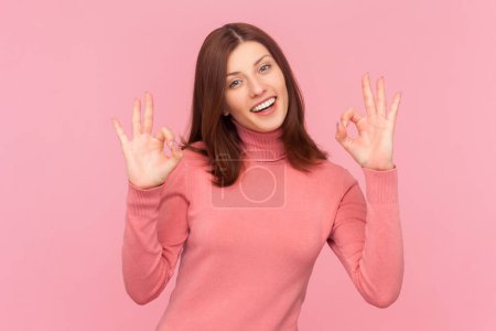 Photo for Joyful woman with brown hair making okay gesture, enjoying life, saying ok, confirms information, its excellent result, wearing rose turtleneck. Indoor studio shot isolated on pink background - Royalty Free Image