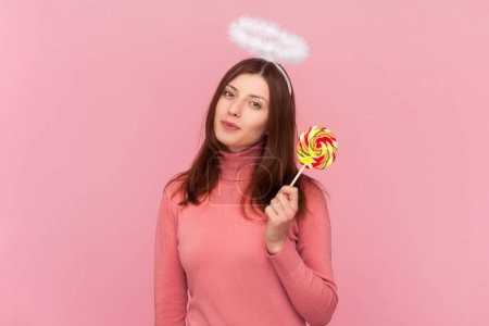 Winsome cute charming woman with brown hair and nimb over head, looking at camera and holding colorful delicious lollipop, wearing rose turtleneck. Indoor studio shot isolated on pink background