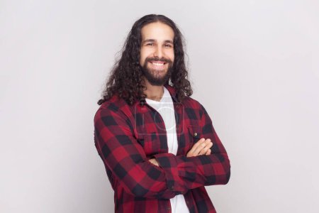 Photo for Portrait of satisfied cheerful handsome bearded man with long curly hair in checkered red shirt looks at camera, keeps hands folded. Indoor studio shot isolated on gray background. - Royalty Free Image